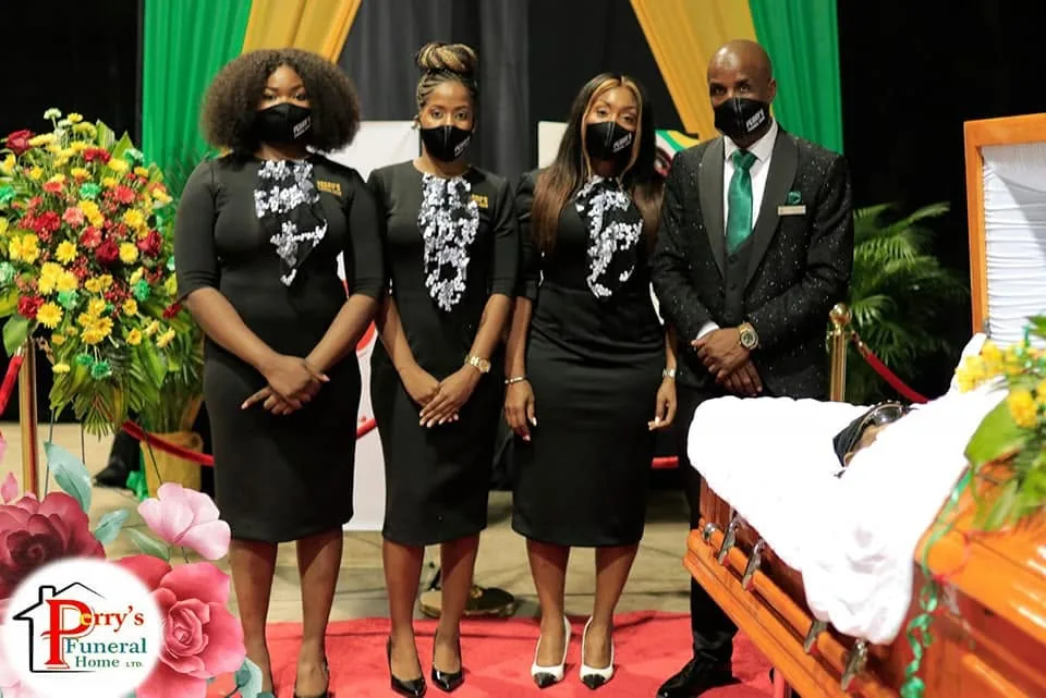 Perry's team standing respectfully beside a casket and floral tributes during a funeral service